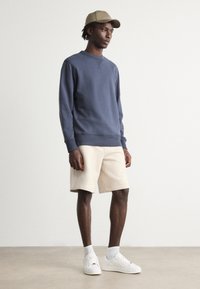Navy sweatshirt with crew neckline, beige shorts, white sneakers, and light-colored socks. Model poses with hands at sides, neutral background.