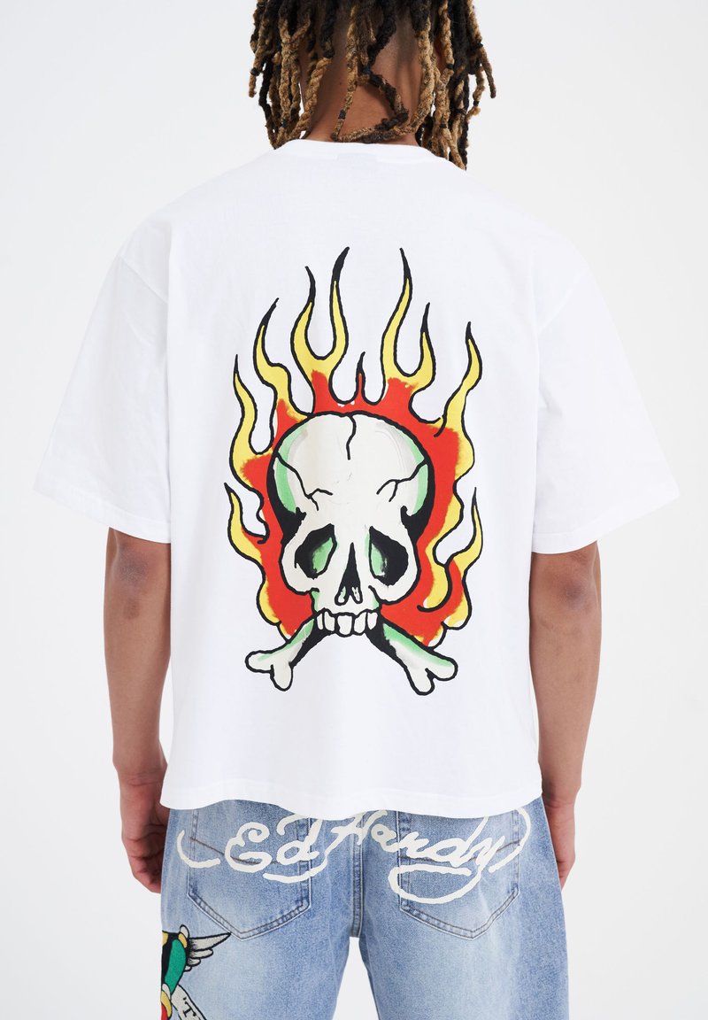 Hvid oversized t-shirt med et farverigt kraniet grafik med flammer på ryggen. T-shirten har en rund halsudskæring og korte ærmer.