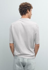 Ljust grå polo t-shirt med mjuk textur, korta ärmar och ribbad krage. Tillsammans med ljus tvättade denimjeans, sedd bakifrån.