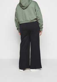 Persona che indossa pantaloni neri a gamba larga, sneakers bianche e una felpa corta verde oliva, in piedi contro uno sfondo bianco uniforme.