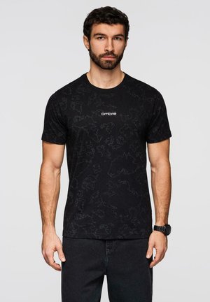 Mann mit dunklem lockigem Haar und Bart, trägt ein schwarzes T-Shirt mit Muster und dem Schriftzug "ombre" sowie eine schwarze Armbanduhr, steht vor einem einfarbigen Hintergrund.
