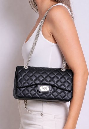 Vrouw in een witte tanktop en broek houdt een zwarte doorgestikte schoudertas vast met een zilveren kettinghengsel en een metalen sluiting.