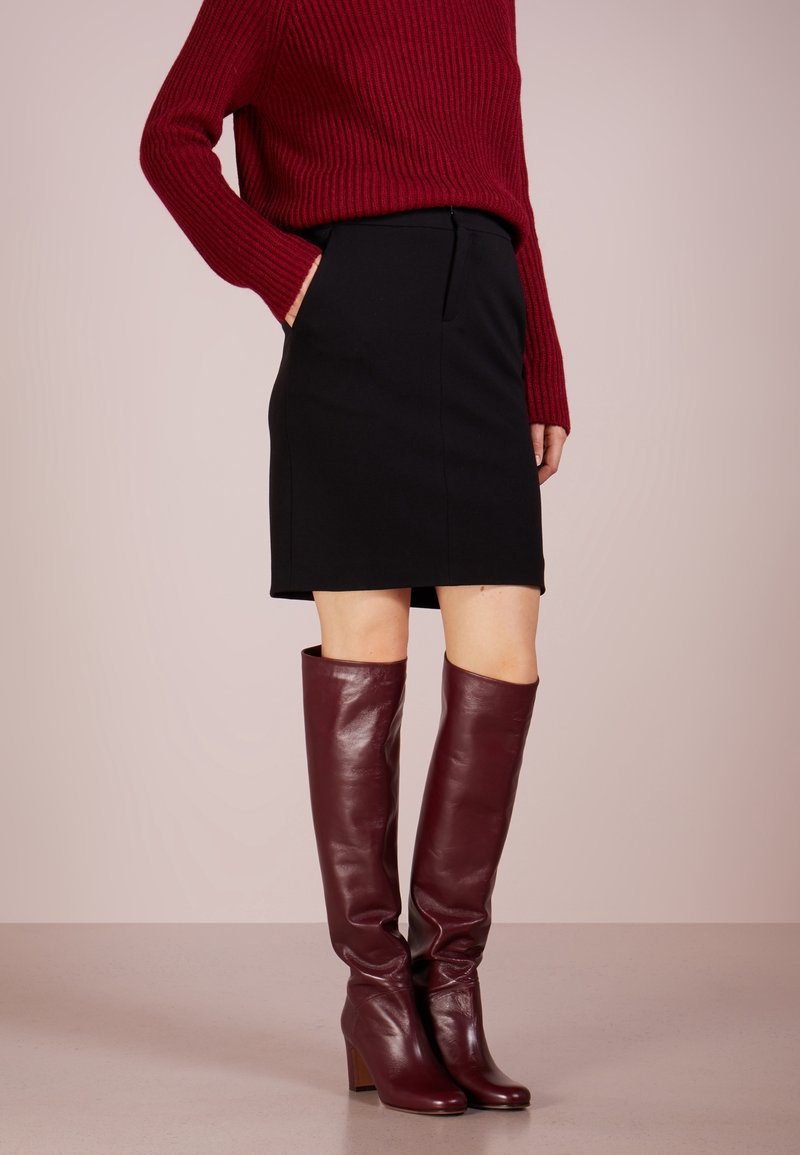 Personne portant un pull rouge côtelé, une jupe noire arrivant aux genoux, et des bottes en cuir rouge foncé montantes jusqu'aux genoux, debout devant un fond uni.