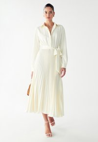 Plissiertes Midikleid aus weichem Elfenbeinmaterial mit einem kragenlosen Oberteil, langen Ärmeln und einem gebundenen Gürtel in der Taille; kombiniert mit Riemchensandalen.