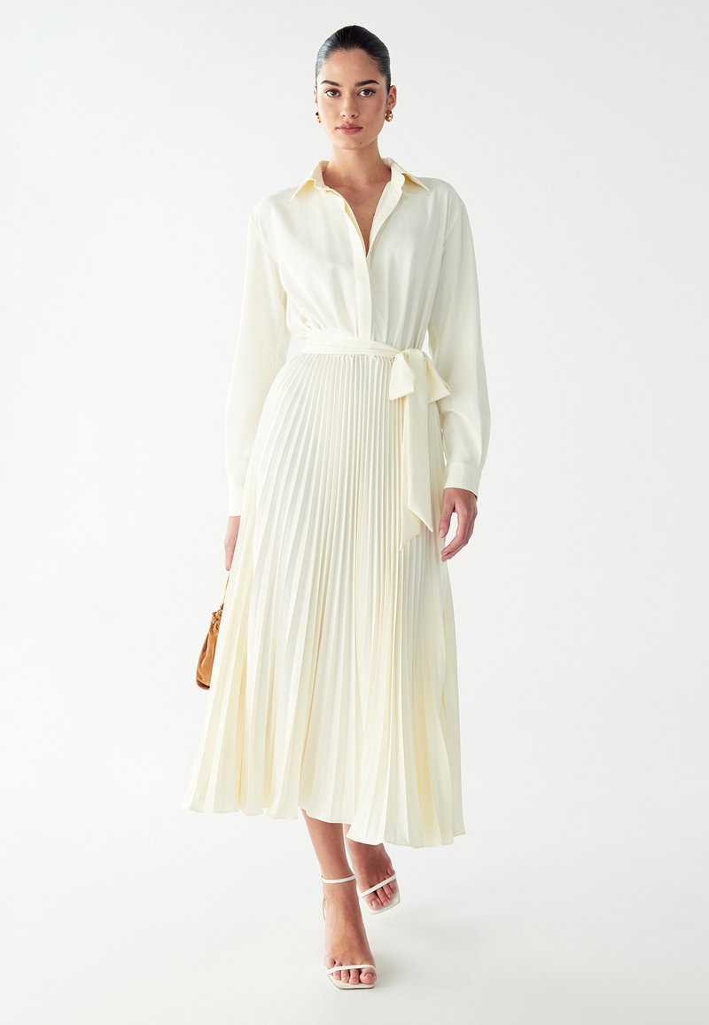 Plissiertes Midikleid aus weichem Elfenbeinmaterial mit einem kragenlosen Oberteil, langen Ärmeln und einem gebundenen Gürtel in der Taille; kombiniert mit Riemchensandalen.
