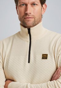 Beige kwart-rits pullover van gestructureerde stof, met een zwarte rits en een klein logo-patch op de borst.
