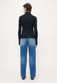 Person with short curly hair wearing a fitted black turtleneck and loose straight-leg blue jeans, standing against a plain background.