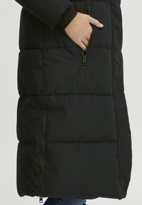 Svart pufferjakke med quiltet design, med sidelommer og lomme foran. Materialet har en glatt, lett glansfull tekstur.