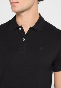 Czarny polo z klasycznym kołnierzykiem, dwoma jasnymi guzikami i haftowanym logo na piersi, wykonany z teksturowanej tkaniny.