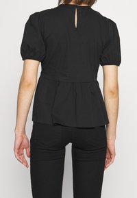 Femme portant un chemisier péplum à manches courtes noir avec une ouverture en keyhole dans le dos et un pantalon noir ajusté, vue de dos.