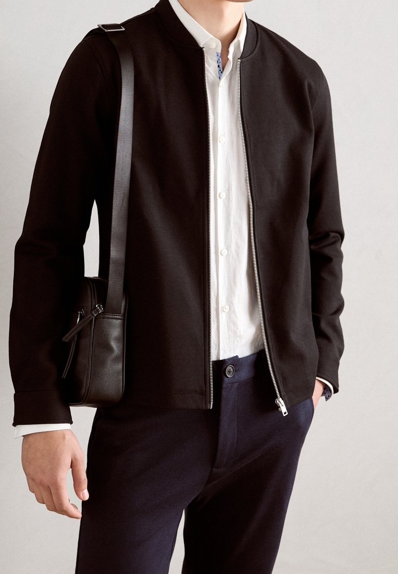 Homme portant une veste zippée noire par-dessus une chemise blanche à boutons, un pantalon bleu marine, et portant un sac noir porté en bandoulière sur un fond clair.