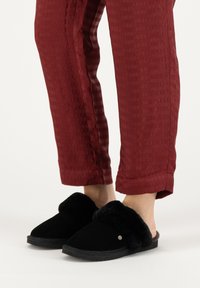 Zwarte pantoffels met een gevoerde voering van bont, met een open achterkant en platte zool, gedragen met rode textuurbroek met een ruitpatroon.