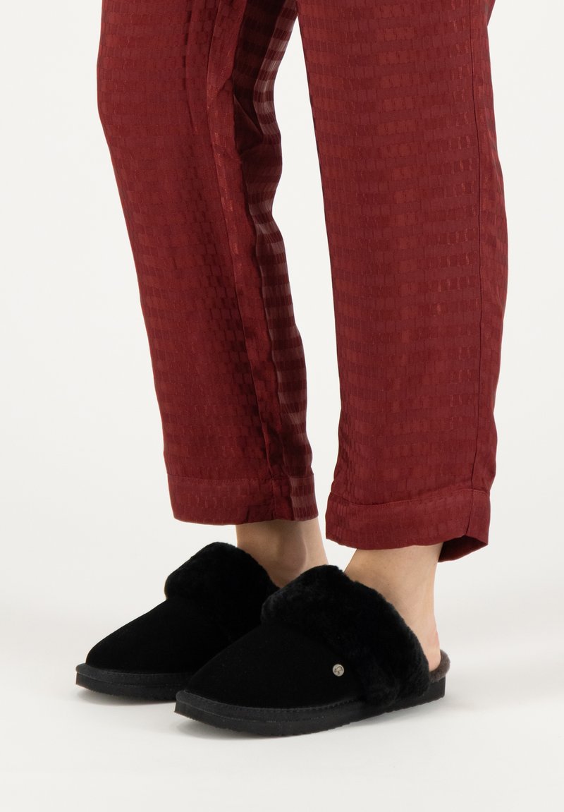 Zwarte pantoffels met een gevoerde voering van bont, met een open achterkant en platte zool, gedragen met rode textuurbroek met een ruitpatroon.