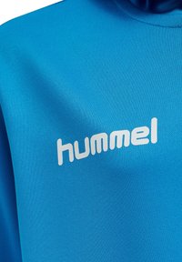 Tessuto blu con logo testuale bianco "hummel" stampato sul petto di un indumento, che mostra la texture del tessuto e i dettagli delle cuciture.