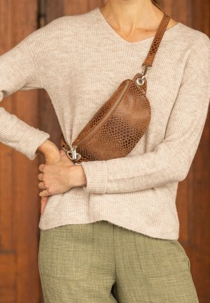 Frau trägt einen beigen Strickpullover und grüne Hose, hält eine braune, strukturelle Leder-Crossbody-Tasche über der Brust.