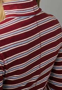 Gros plan sur un pull à col roulé côtelé bordeaux avec des rayures horizontales blanches et bleu clair porté par une personne.