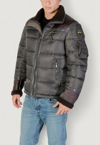 Uomo che indossa una giacca imbottita grigio scuro con polsini e colletto in shearling marrone, tasche con zip e jeans azzurri chiari, su sfondo neutro.