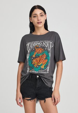 T-shirt in cotone grigio con una grafica colorata di tigri e testo, abbinata a pantaloni corti in denim neri strappati per un look casual.