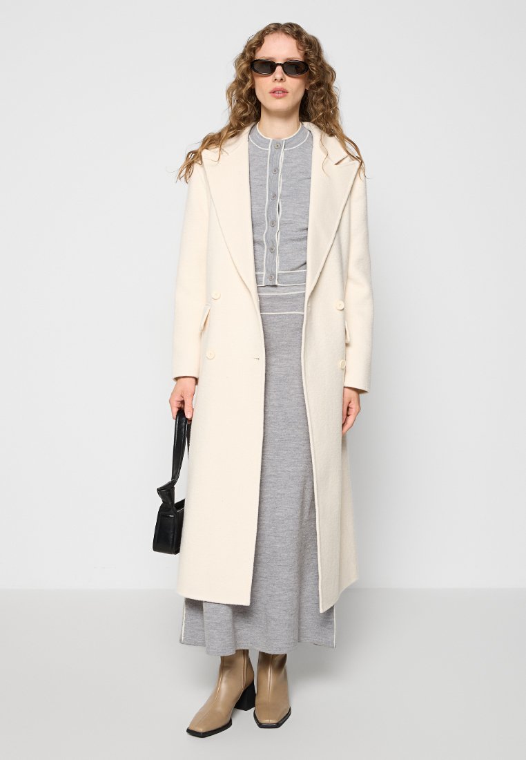 Langer cremefarbener Wollmantel mit großen Revers, kombiniert mit einem grauen Strickkleid. Beigefarbene Knöchelstiefel und eine kleine schwarze Handtasche. Die Accessoires umfassen Sonnenbrillen.
