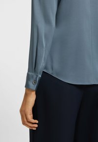 Zachte, gedempte blauwe blouse met knoopdetails bij de pols. Gecombineerd met donkere, op maat gemaakte broek, die een gladde textuur en scherp ontwerp tentoonstelt.