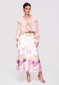 Femme en blouse rose clair à volants, jupe midi blanche à fleurs avec boutons et ceinture beige, portant des sandales à talons hauts couleur peau, debout.