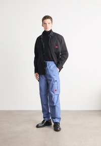 Casaco preto de botões sobre um pulôver preto, combinado com calças cargo azuis com bolsos e apliques, e sapatos pretos.
