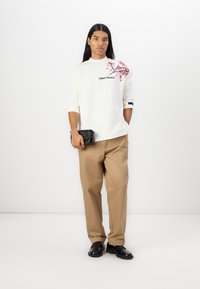 Sweatshirt blanc avec imprimé floral rose et logo ; pantalon beige ; le mannequin tient un petit sac noir ; chaussures noires. Style simple et décontracté.