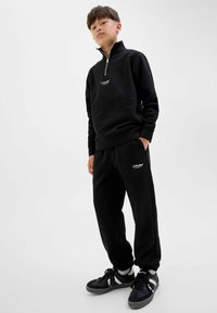 Svart zip-nack sweatshirt och matchande sweatpants, båda med ett mjukt tyg, med broderade logotyper på bröstet och låret. Athletiska skor ingår.