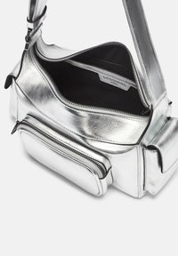 Srebrna metaliczna torba na ramię o smukłym designie, z zamkniętą na zamek główną komorą i przednią kieszenią, wnętrze jest czarne.