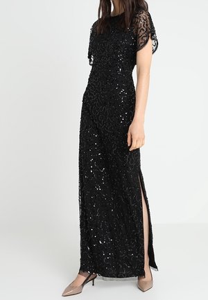 Robe noire longue jusqu'au sol ornée de sequins, avec manches courtes transparentes et fente latérale, portée avec des escarpins beiges à bout pointu.