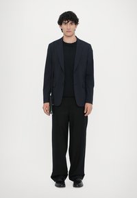 Sort blazer over en sort sweater, parret med brede sorte bukser og sorte sko. Personen holder en lille sort taske.