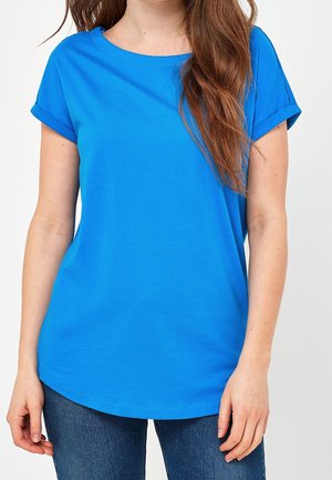 Vrouw draagt een effen felblauw T-shirt met korte mouwen en donkerblauwe spijkerbroek, staand tegen een witte achtergrond.