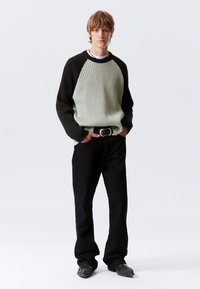 Knitted sweater in light green and black with ribbed texture and raglan sleeves, paired with black pants and a black belt, set on a plain background.
