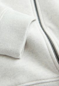 Gros plan sur le poignet d’un sweat-shirt gris clair et une fermeture éclair argentée sur un tissu doux côtelé.