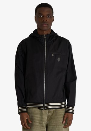 Giovane uomo con giacca nera con zip, cappuccio e polsini a righe, in piedi davanti a uno sfondo chiaro e uniforme.