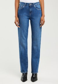 Osoba ubrana w niebieskie jeansy o prostych nogawkach z przednimi kieszeniami oraz czarne botki, stojąca na jednolitym tle.