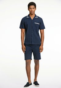 Conjunto de camisa de manga corta y pantalones cortos de color marino con tejido texturizado; la camisa presenta un cuello blanco, un bolsillo y botones en la parte delantera; combinado con zapatos oscuros de slip-on.