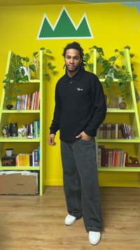 Μαύρο μπλουζάκι πόλο μακρυμάνικο με διακριτικό μοτίβο, συνδυασμένο με σκούρο γκρι παντελόνι φαρδιάς γραμμής και λευκά αθλητικά παπούτσια, στημένα μπροστά από έναν κίτρινο τοίχο.