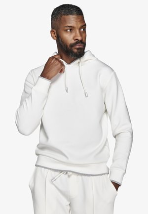 Partas mies valkoisessa hupparissa ja siihen sopivissa valkoisissa housuissa, pitelee hupparin nyöriä kaulan lähellä ja katsoo sivulle.