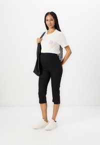 T-shirt blanche avec un graphique rose, associée à un pantalon noir court et des baskets blanches. Le modèle porte un sac noir, se tenant devant un fond uni.