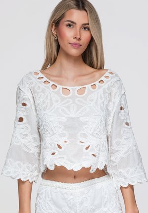 Junge Frau mit langen blonden Haaren, die ein weiß besticktes Croptop mit wellenförmigen Rändern und passenden weißen Unterteilen trägt.