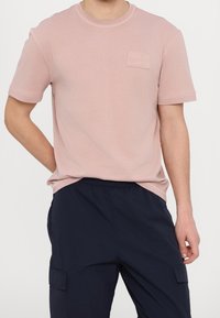 Lichtroze katoenen t-shirt met korte mouwen, ronde hals en een klein reliëf logo-label op de linkerborst. Zachte textuur, casual pasvorm.