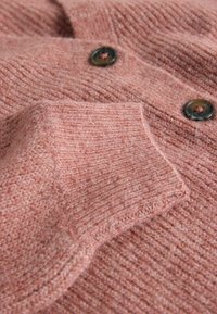 Hieman teksturoitu vaaleanpunainen neulepaita, jossa on kaksi tummaa nappia ja käänteinen hihadetaili, esittelee pehmeää ja mukavaa materiaalia.