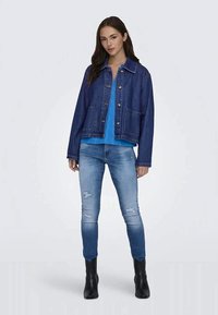Jovem mulher vestindo uma jaqueta de denim azul escuro, blusa azul brilhante, jeans skinny desbotados com rasgos e botas pretas até o tornozelo, posando contra um fundo simples.