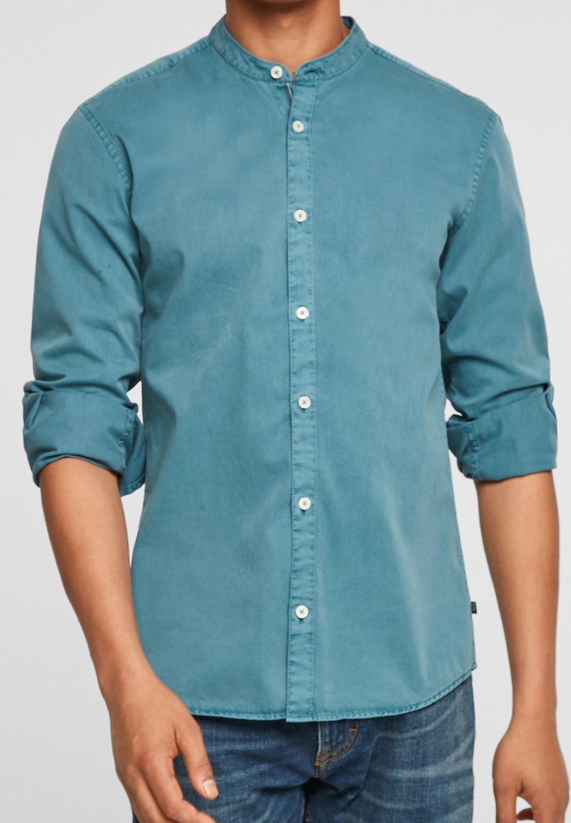 Chemise boutonnée couleur sarcelle avec un col montant, des manches longues retroussées et une patte de boutonnage avant avec des boutons blancs ; tissu en coton à la texture lisse.