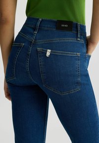 Hubené modré džíny s vysokým pasem, kontrastním prošíváním a stříbrným kovovým detailem na zadní kapse. Na pásku je štítek s nápisem "LIUJO."