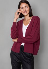 Burgundy knit cardigan with dropped shoulders, ribbed cuffs and hem, layered over a white top. Paired with black drawstring pants.