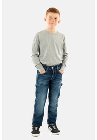 Camisa gris de manga larga, jeans de mezclilla azules y zapatillas negras. Los jeans tienen un corte recto y detalles de costura visibles. Manos en los bolsillos.