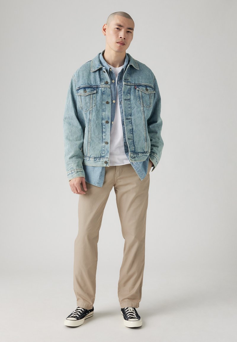 Veste en denim bleu clair avec deux poches poitrine, associée à un t-shirt blanc, un pantalon beige et des baskets noir et blanc.