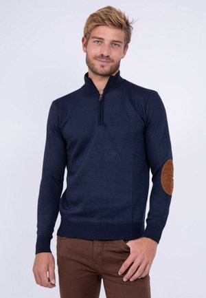 Homme aux cheveux châtain clair portant un pull bleu marine à fermeture éclair quart de zip avec des coudières marron et un pantalon marron, debout devant un fond clair.